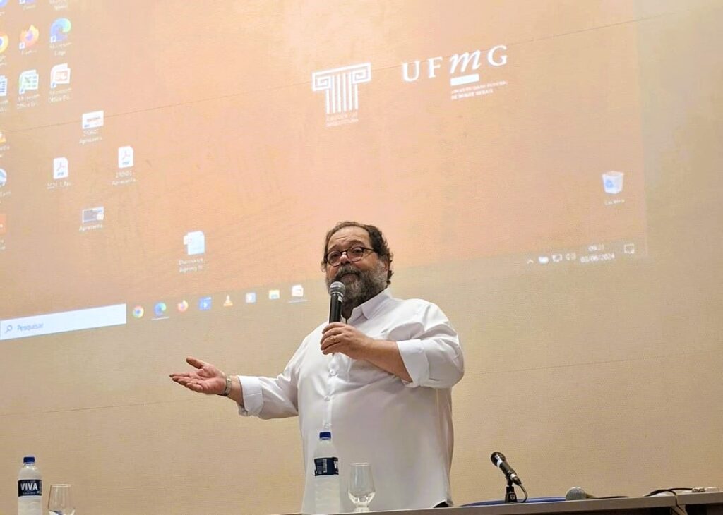 A foto mostra o professor Maurício Campomori, homem branco, forte, explanando. AO fundo há uma projeção da tela inicial de um computador, onde aparece o logo da Escola de Arquitetura e o da UFMG, em um fundo em tom de cor bege.