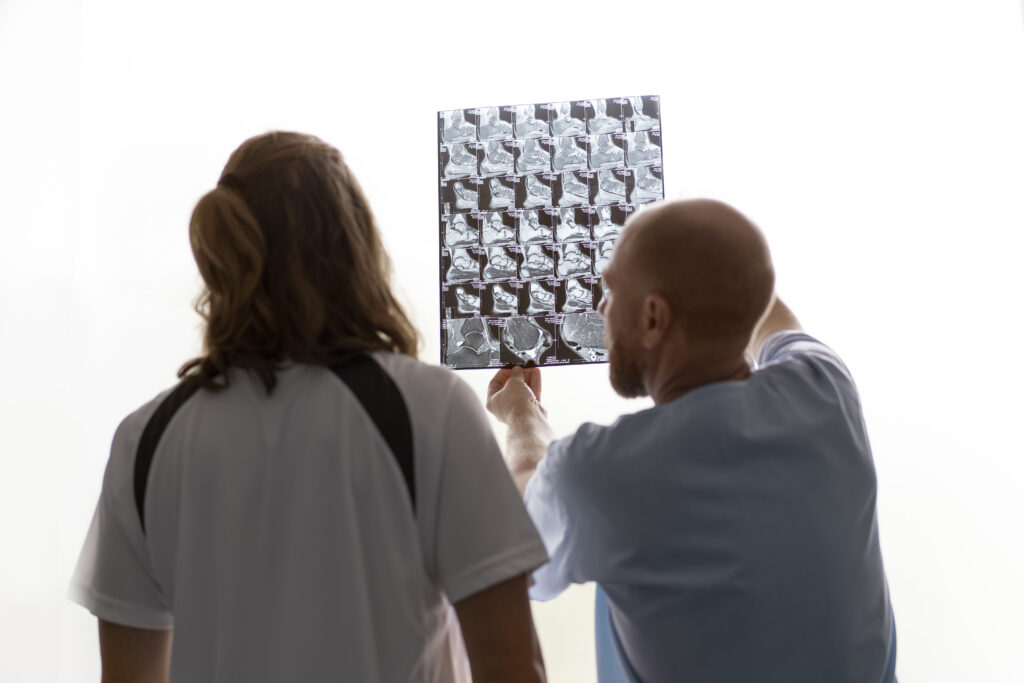 Imagem ilustrativa mostra um homem e uma mulher aparentando serem médicos, observando um exame de imagens do cérebro. O homem, branco, com cabelo curto e barba, está explicando algo sobre a imagem para a mulher, branca, com cabelo longo e solto, que observa atentamente.